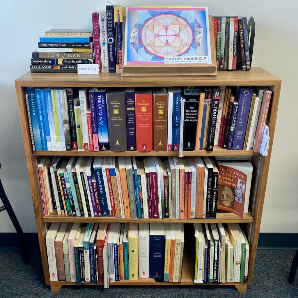 A wooden bookshelf full of books. Loose books as well as a binder on a stand sit on the top.