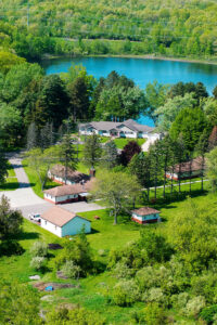 An aerial view of Maryville Retreat Center. The view is sunny and full of lush green foliage. A small lake is visible beyond the grouping of several buildings.